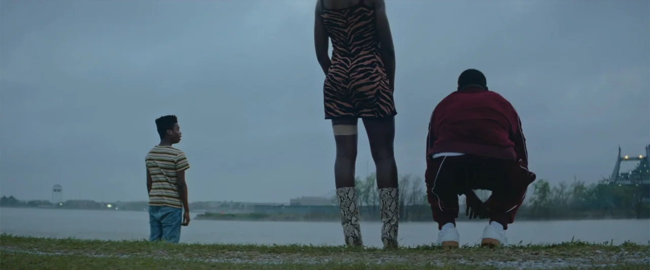 Queen & Slim (2019) movie still: fullbody — A flat riverbank under a heavy overcast sky with a distant industrial water tower. Three people stand or…