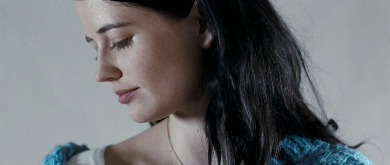 Womb (2010) movie still: closeup — A woman stands indoors against a plain light gray wall. She looks downward with her head turned partially toward…