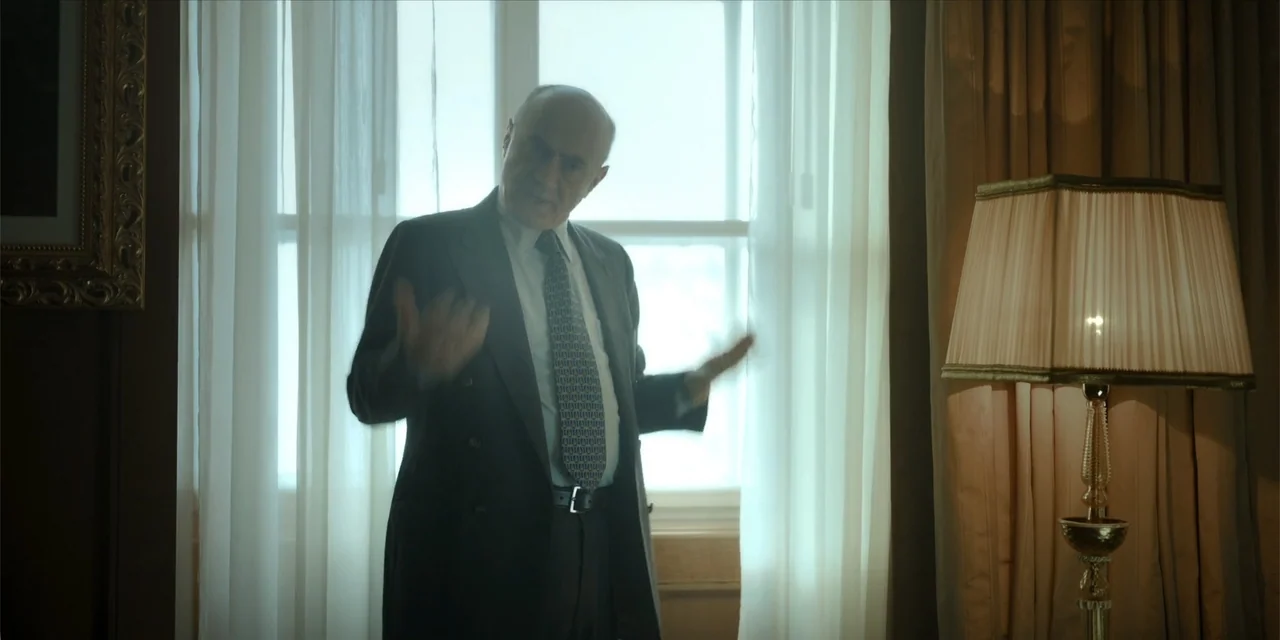 Oslo (2021) movie still: medium — A well-lit office interior features a large window covered by light sheer curtains. An elderly man in a dark suit…