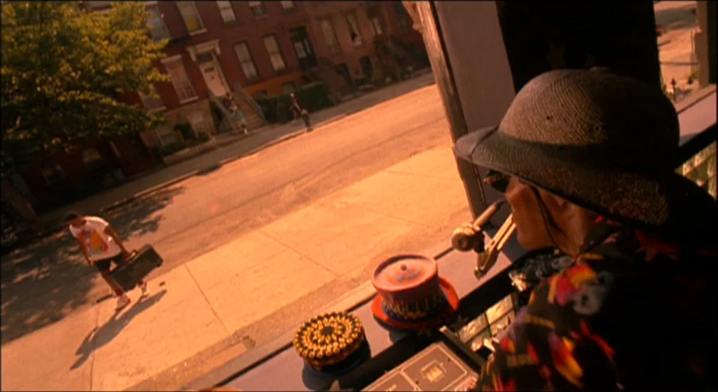 Do The Right Thing (1989) movie still: medium — An older man stands inside a store looking out a window onto a city street. A young man carries a…