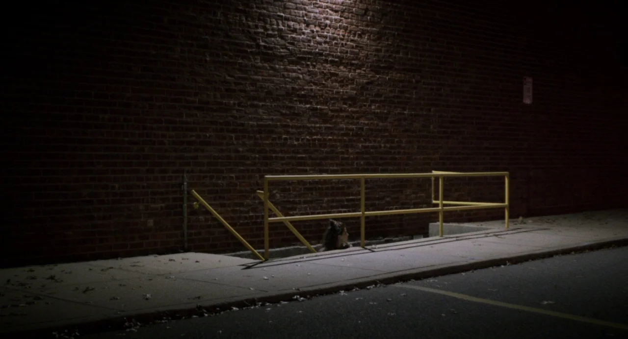 Happer’s Comet (2022) movie still: wide — A night scene shows a dark brick wall bordering a paved sidewalk at street level. A woman emerges from a…