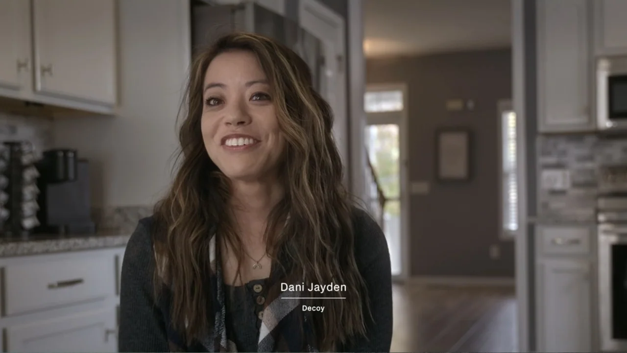 Predators (2010) movie still: medium — A modern kitchen with white cabinetry and a partially visible blurred background hallway. A woman sits facing…