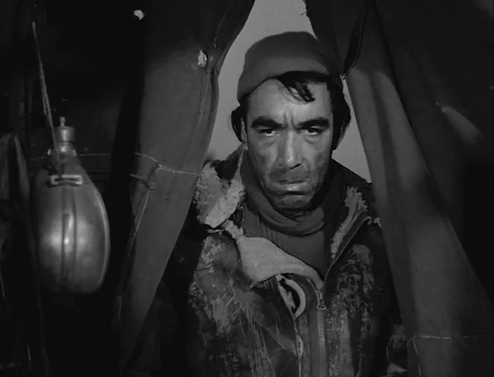 La Strada (1954) movie still: closeup — A man stands framed between two dark, heavy curtains in a field environment. He looks directly ahead with a…