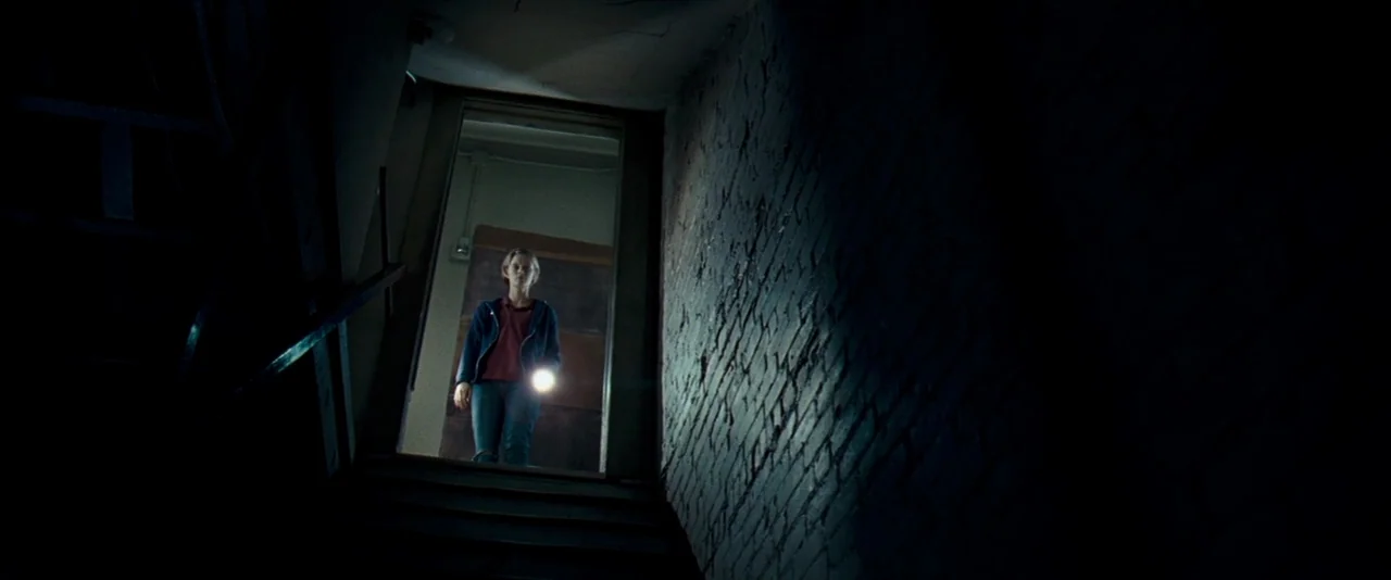 The Innkeepers (2011) movie still: medium — A dark stairwell constructed of uneven brick walls leads up to an illuminated doorway. A young woman…