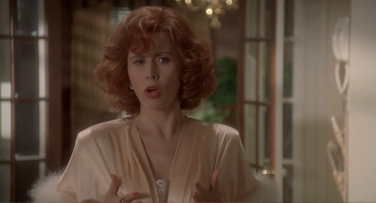 Fatal Instinct (1993) movie still: medium — A well-lit indoor space featuring glass panel doors and soft home decor elements. A woman with auburn…