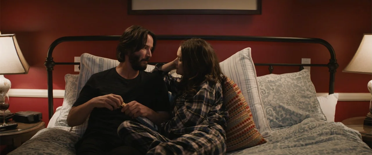 Destination Wedding (2018) movie still: medium — A dimly lit bedroom features deep red walls and a wrought iron bed frame. A man and a woman sit…