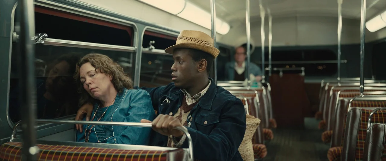 Empire of Light (2022) movie still: medium — The interior of a vintage bus features patterned plaid seats and metal poles. A man with a fedora sits…