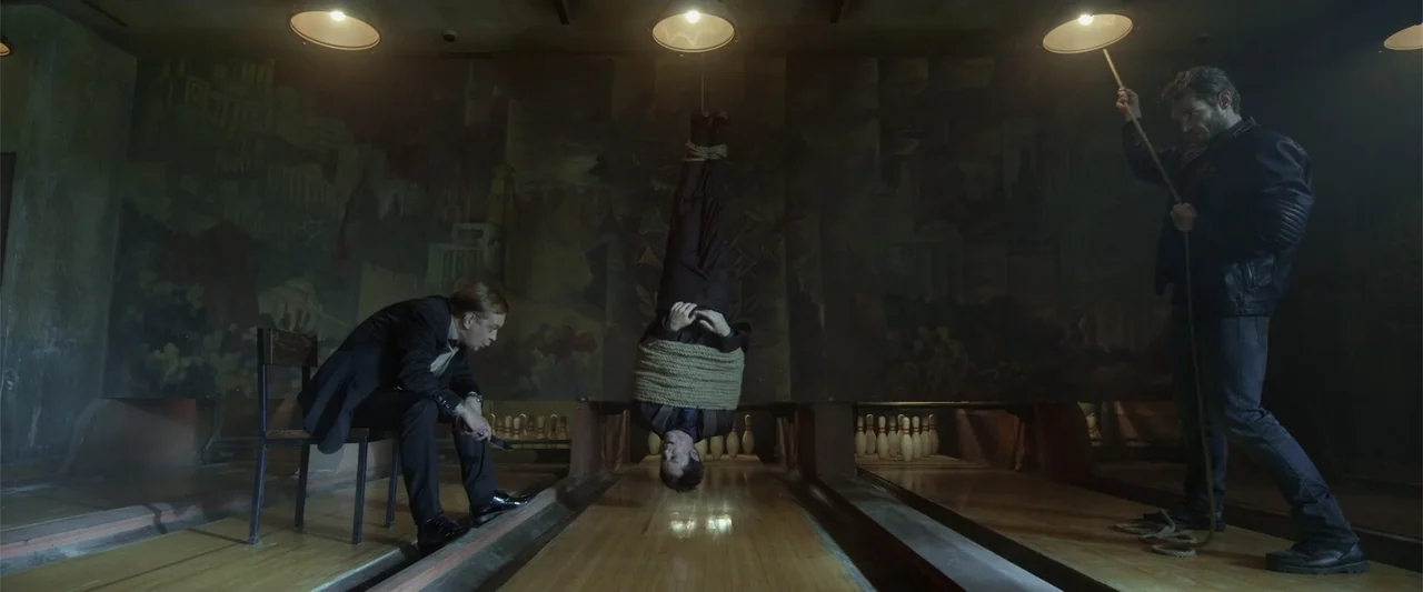 Skiptrace (2016) movie still: wide — A dark, dimly lit venue features weathered wooden lanes and a mural on the wall. Three men interact while one…