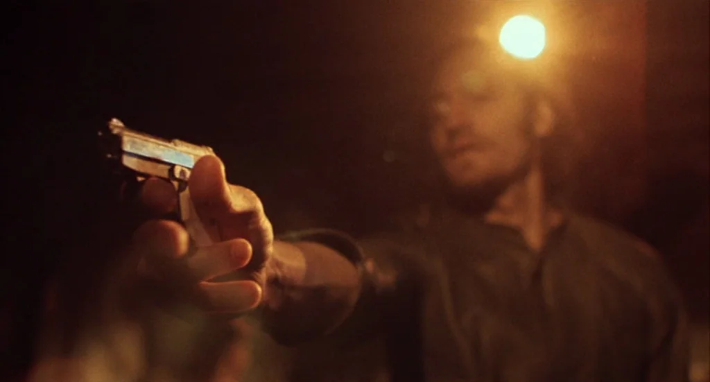 Buffalo ’66 (1998) movie still: medium — A dark outdoor space illuminated by a single bright overhead light source. A man extends his arm while…