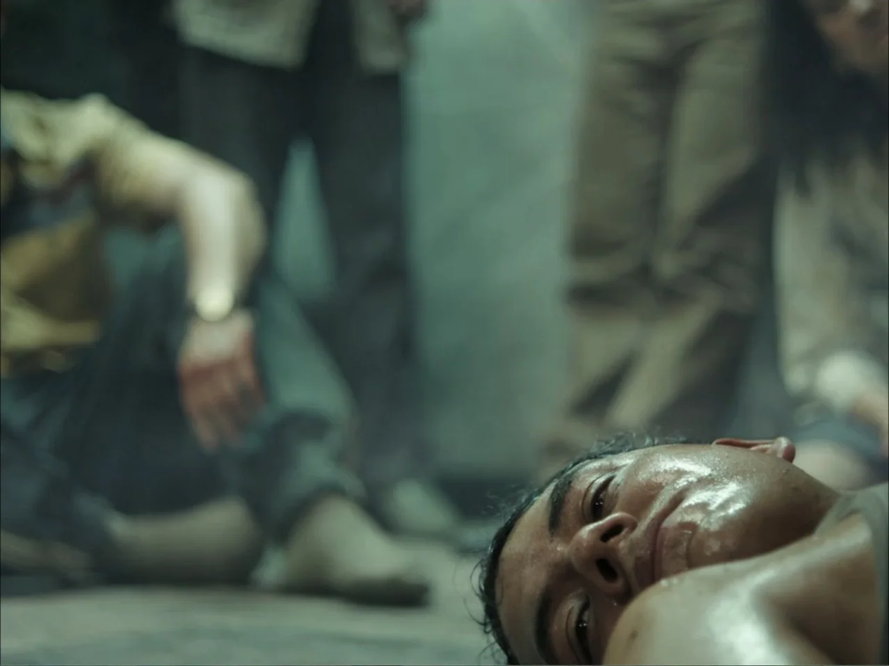 November (2025) movie still: closeup — A dimly lit industrial room with stone-like walls and several people standing in background. A man lies on…