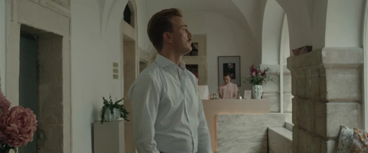 Peacock (2010) movie still: medium — A stone hallway with arched ceilings leads toward a small service reception counter. The man in the foreground…