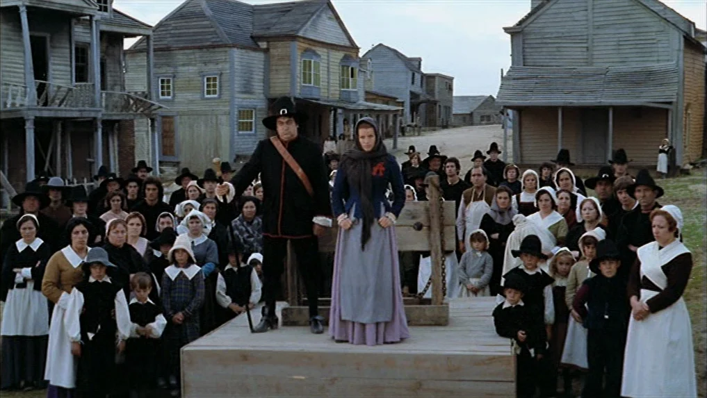 The Scarlet Letter (1973) movie still: wide — A group of villagers gather in a town square with wooden buildings in background. A man and a woman…