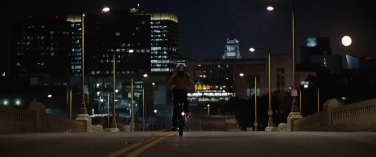 500 Days of Summer (2009) movie still: wide — A night scene shows a child riding a bicycle across a bridge at night. In the background, the…
