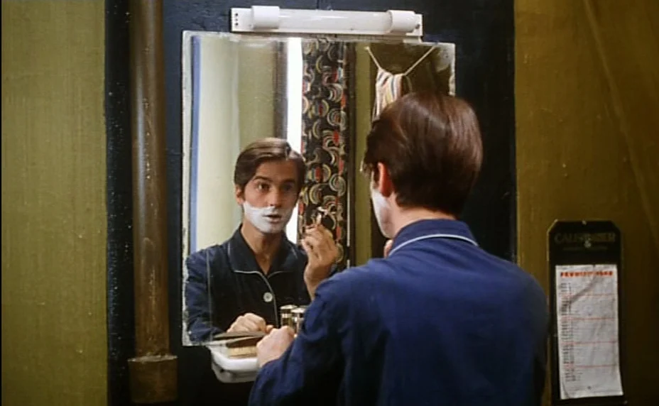 Stolen Kisses (1968) movie still: medium — A cramped bathroom features dark green walls and a wall-mounted rectangular mirror. A man stands before…