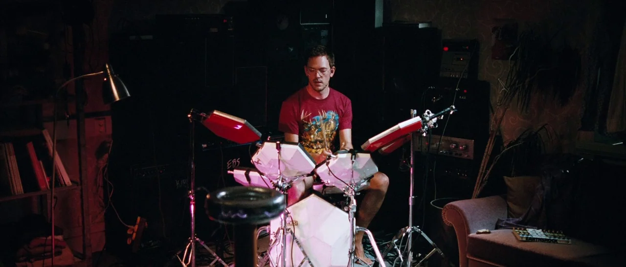 Sound of Noise (2010) movie still: medium — A dimly lit room filled with electronic musical equipment and music storage. A young man sits on a stool…