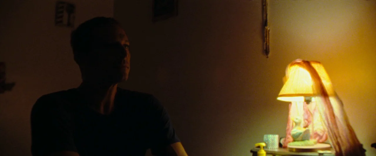 Red Rocket (2021) movie still: medium — A dark space features a man in silhouette sitting beside a small table lamp. The lamp, which has a…