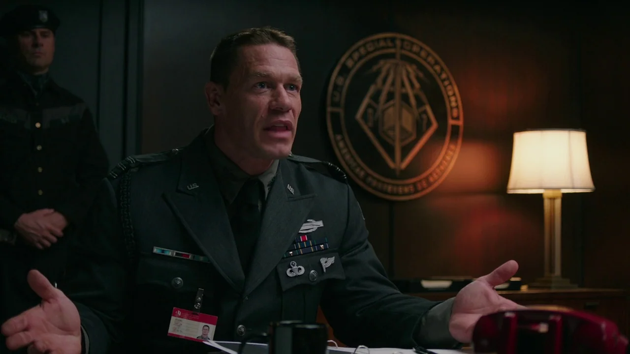 Bumblebee (2018) movie still: medium — An office with a wall-mounted U.S. Special Operations emblem and dim lighting. A military officer sits at a…