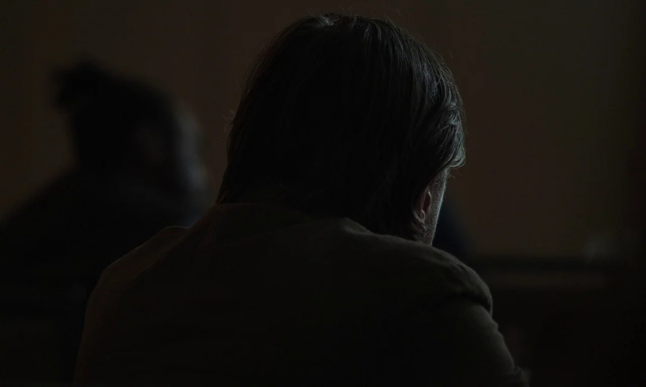 Resurrection (2022) movie still: medium — A dark, dimly lit room with undefined walls and deep shadows. A man sits facing away from the camera,…