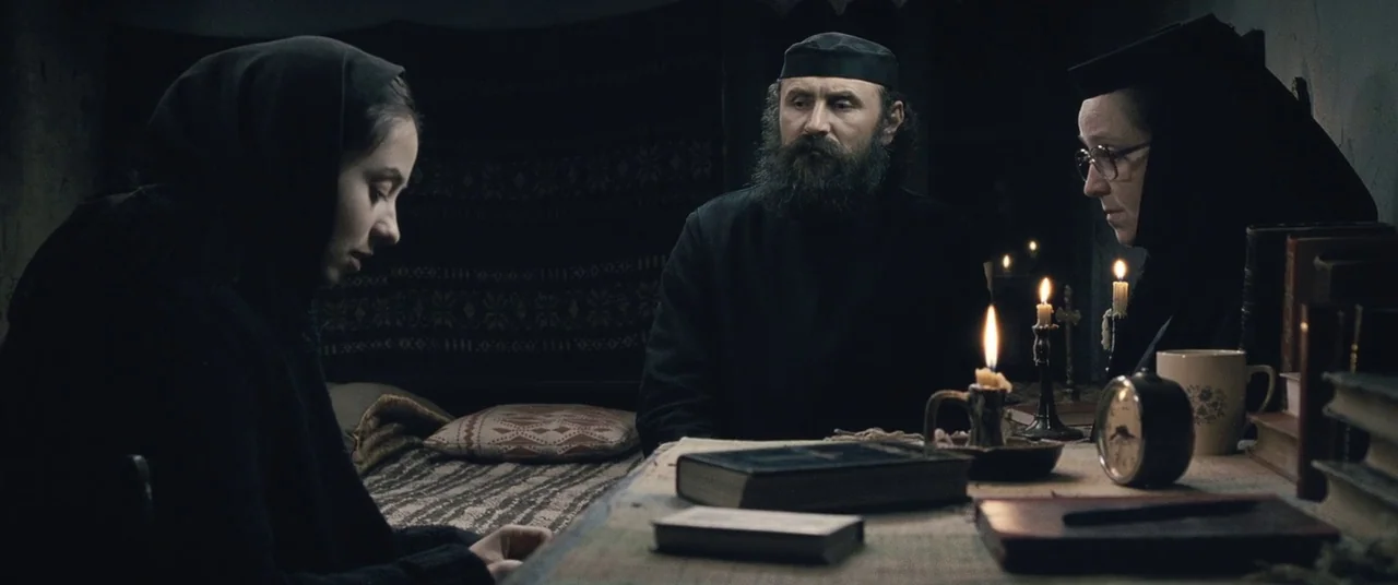 Beyond the Hills (2012) movie still: medium — A dimly lit room features a wooden table scattered with religious books and candles. Three people are…