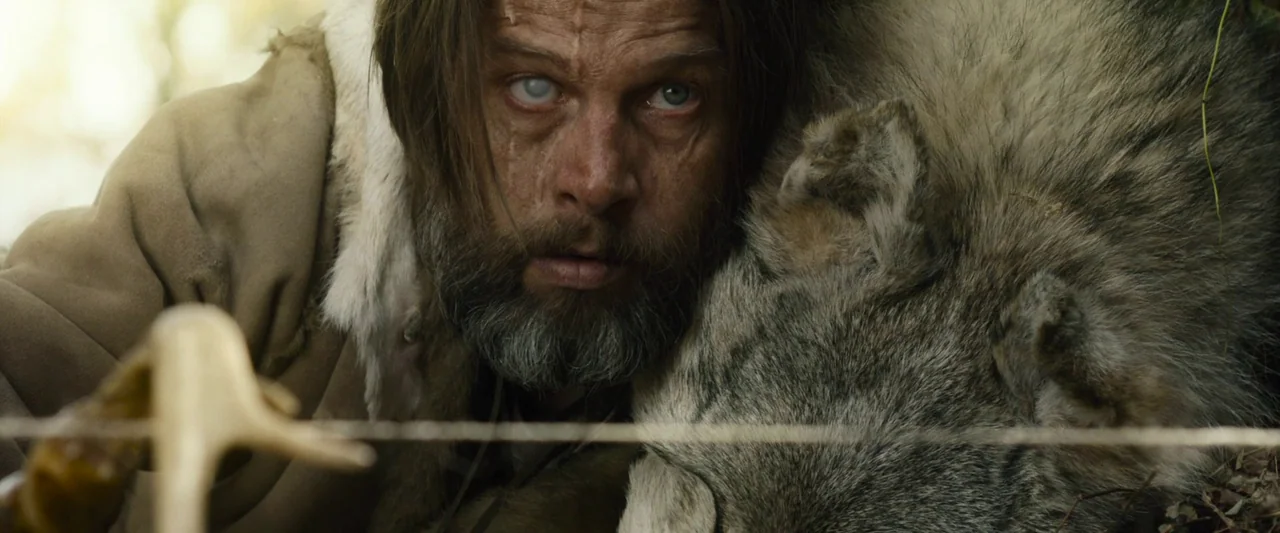Wildling (2018) movie still: closeup — A man lies closely beside a large, gray wolf in an outdoor environment. He wears tattered fur clothing and…