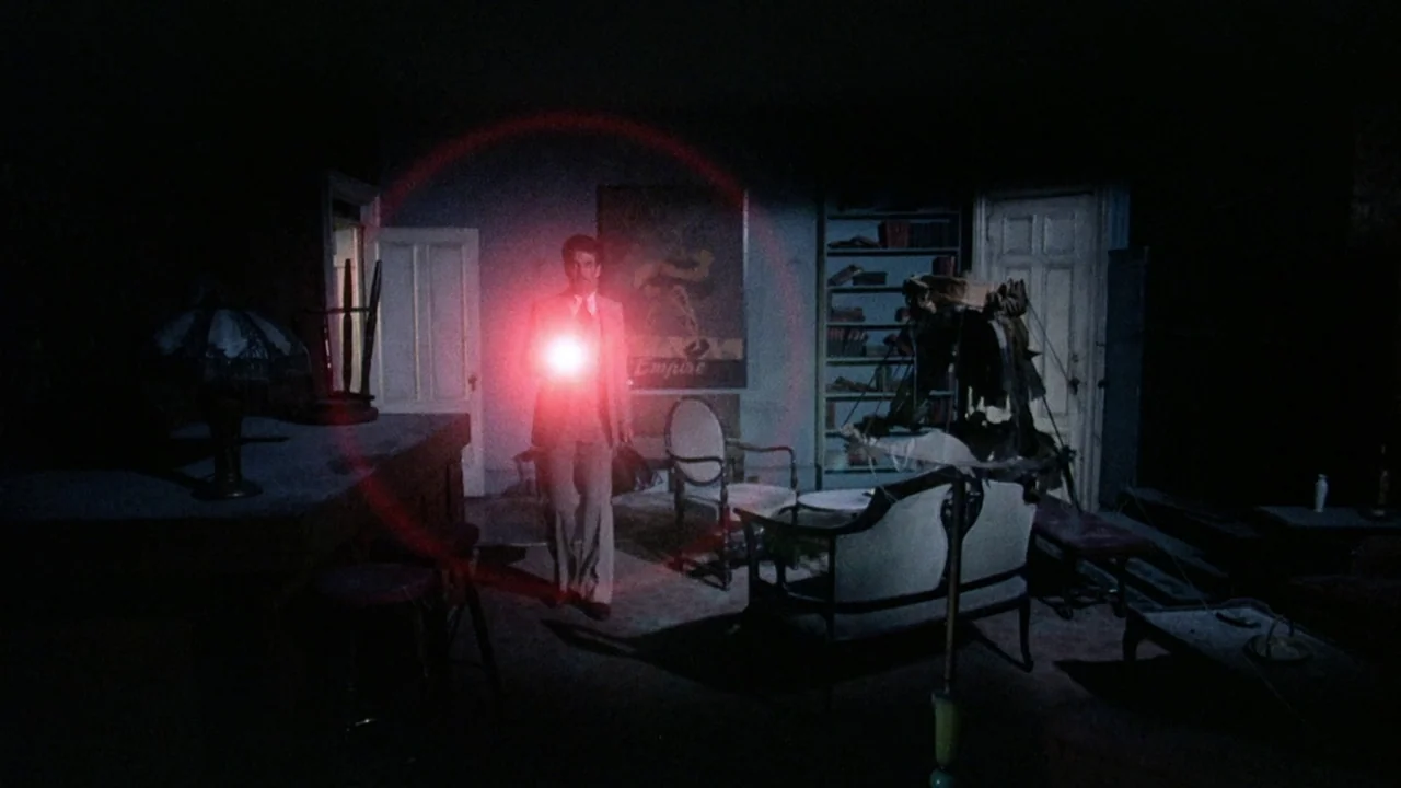 The Sentinel (1977) movie still: fullbody — A dark, cluttered room with aged furniture and a visible poster on the wall. A man stands in the center…