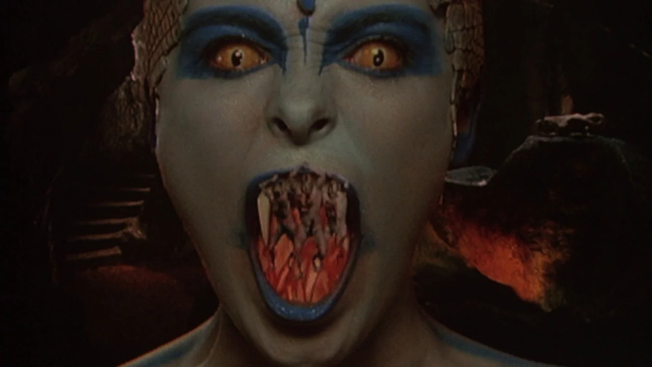 The Lair of the White Worm (1988) movie still: closeup — A close-up of a blue-skinned being against a dark, rocky background. The person opens their…