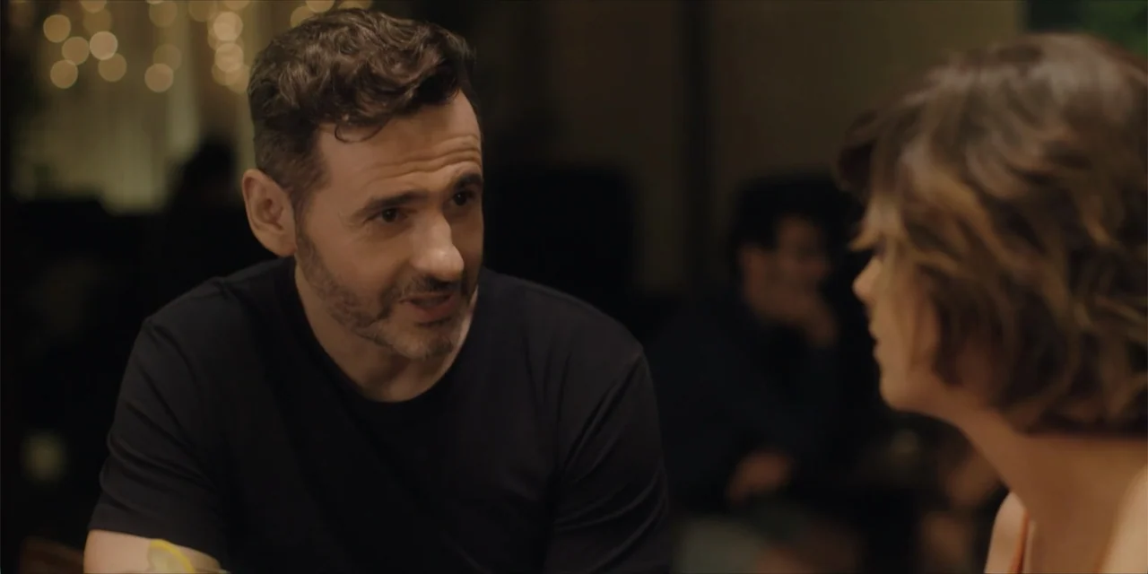 Instant Love (2020) movie still: medium — A dimly lit indoor space with blurred golden bokeh lights in the background. A man faces a woman in a…