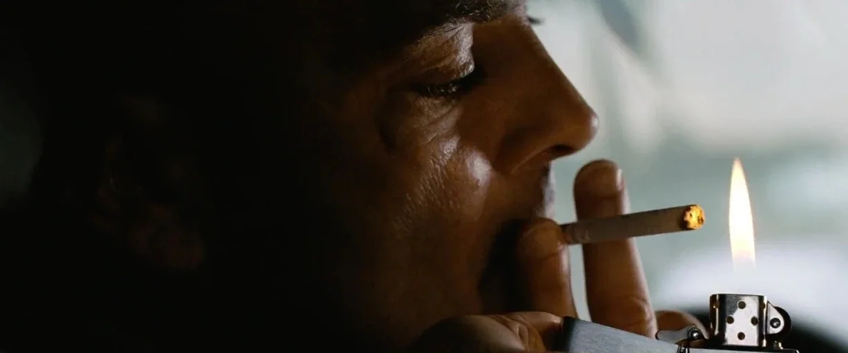 The International (2009) movie still: closeup — A dark vehicle interior with the subject in profile near a blurred window. A man holds a lit…