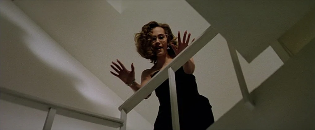 Anguish (1987) movie still: medium — A woman stands behind a minimalist white metal railing of an indoor staircase. She looks downward with her arms…