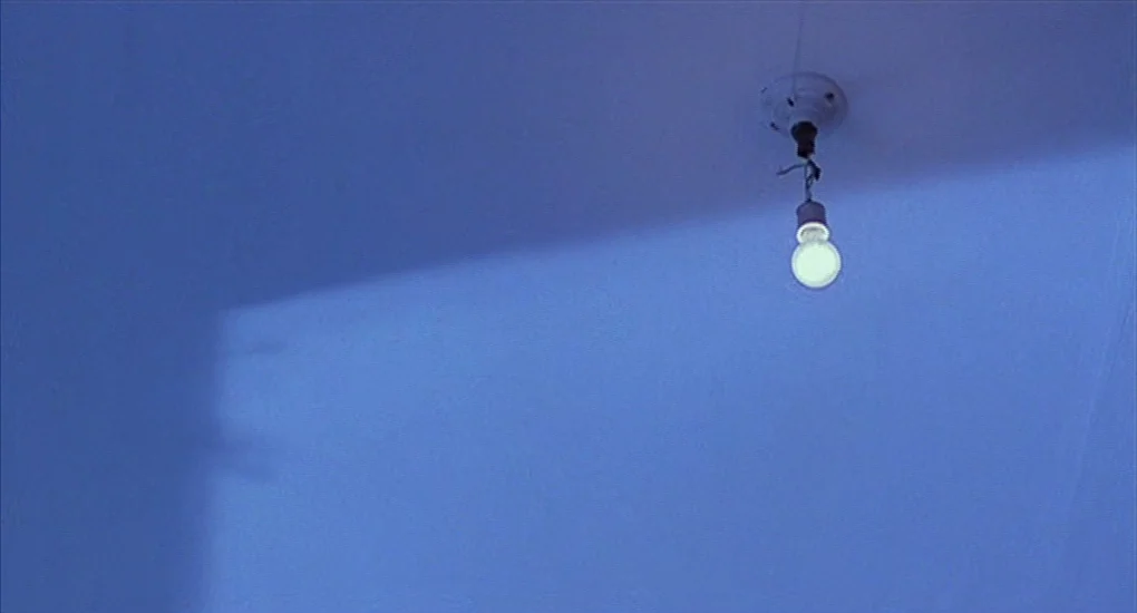 Exotica (1994) movie still: establishing — A plain blue ceiling features a single bare light bulb hanging on a wire. A crisp diagonal shadow cuts…