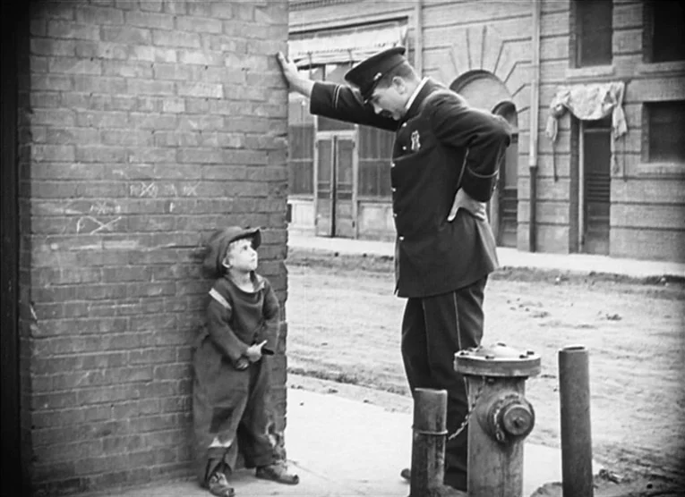 The Kid (1921) movie still: fullbody — A quiet sidewalk bordered by an old brick wall and a distant building facade. A young boy in oversized…