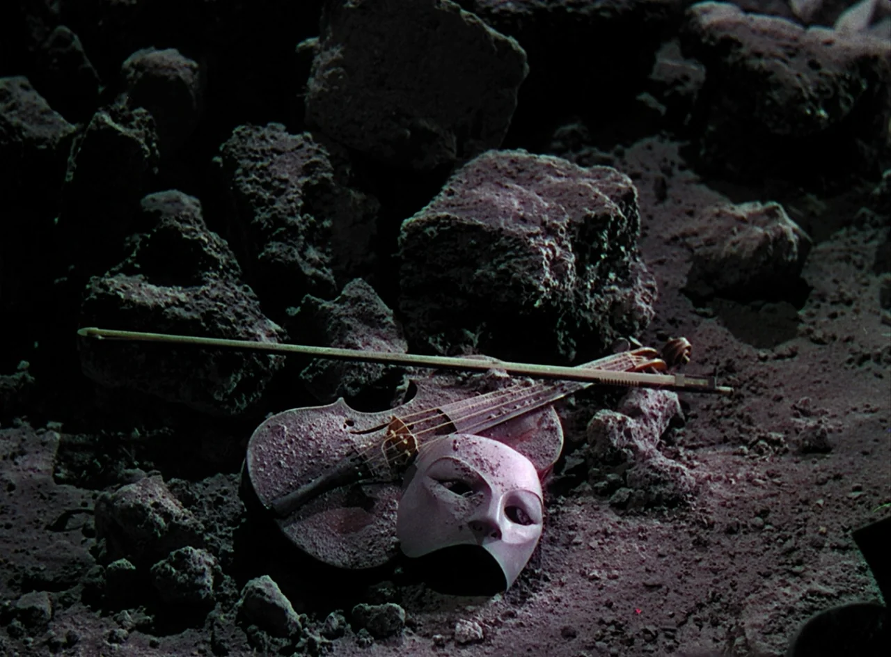 Phantom of the Opera (1943) movie still: closeup — A violin and a smooth white theatrical mask rest on a bed of dark rocks. The instruments are…