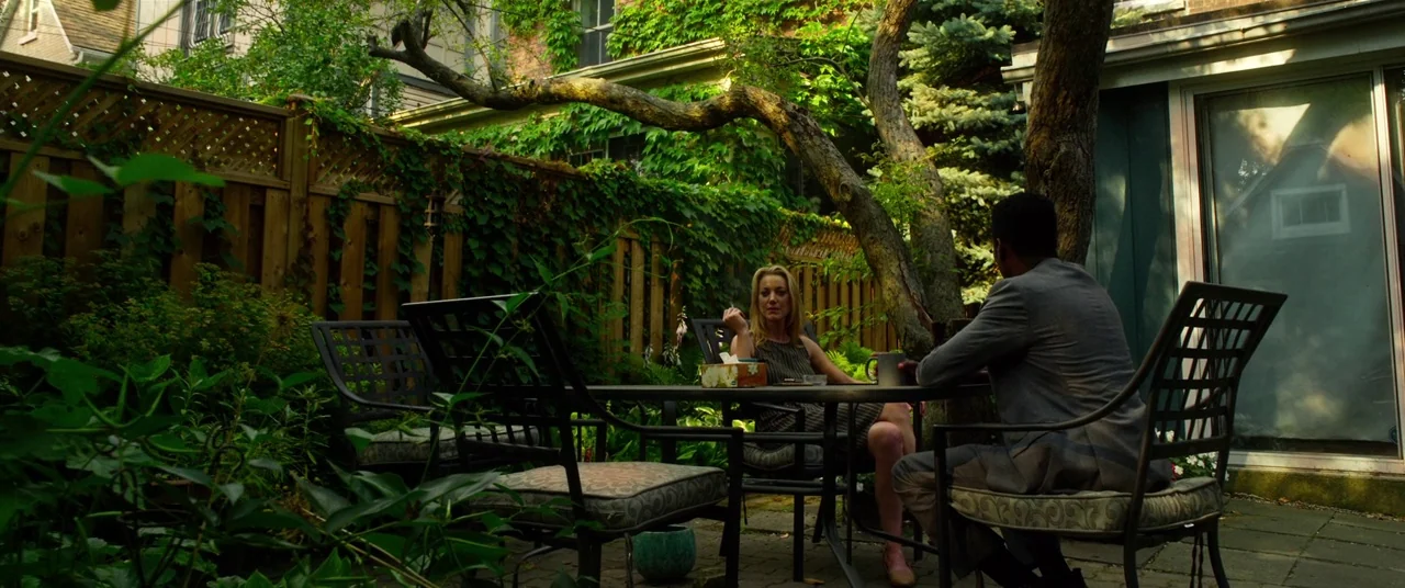 Spiral (2021) movie still: wide — A residential patio surrounded by lush climbing ivy and mature trees on a sunny day. Two individuals sit at a…