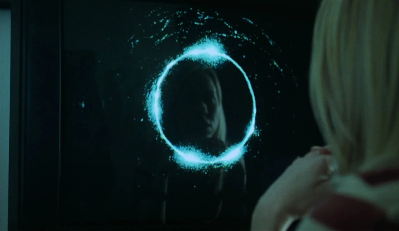 The Ring (2002) movie still: medium — A dimly lit interior space features a glowing blue ring on a screen. A woman with blonde hair watches the…