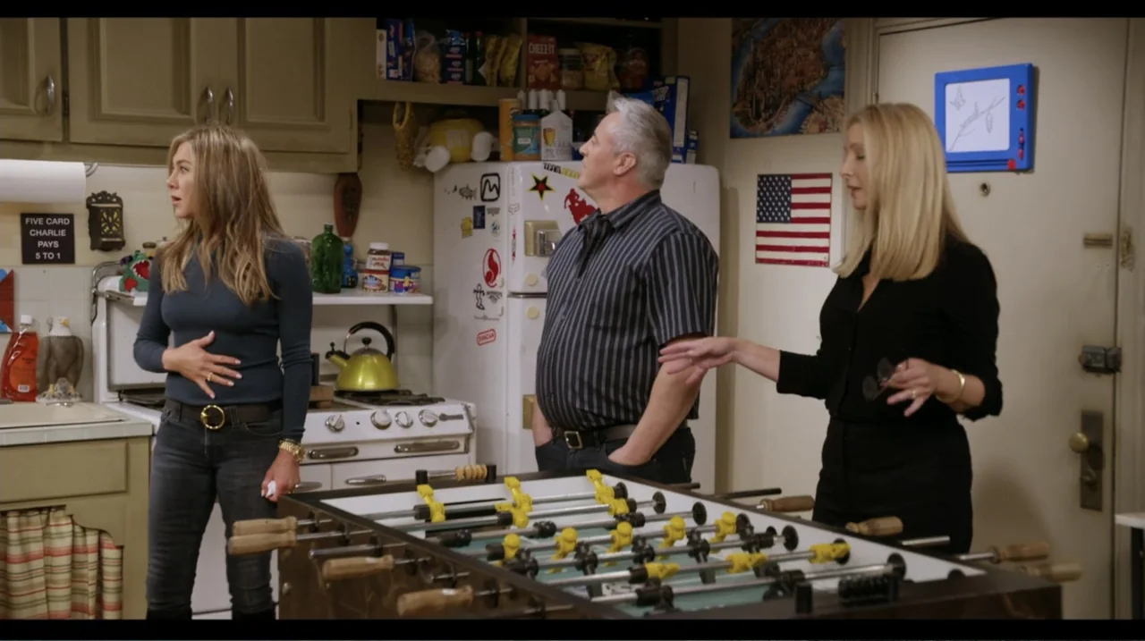 Friends: The Reunion (2021) movie still: fullbody — A cluttered apartment kitchen features a foosball table in the foreground near cabinets. Three…