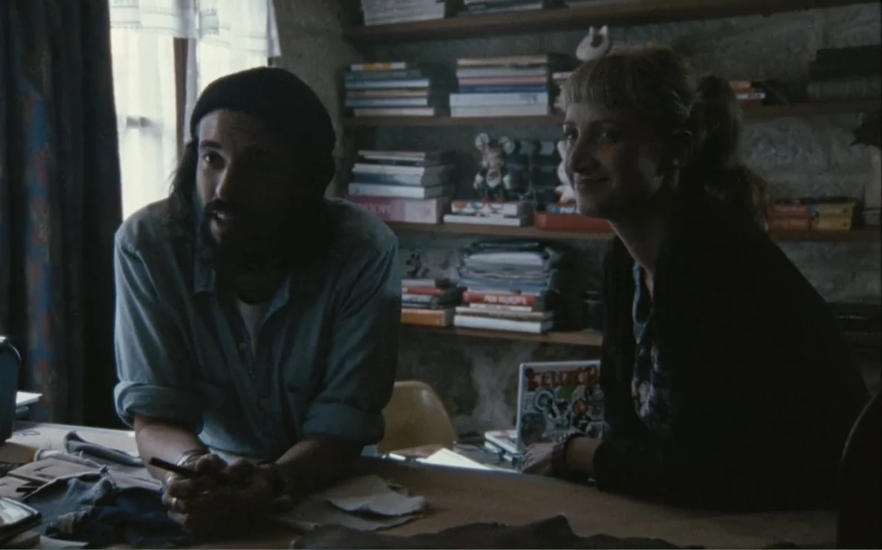 The Greasy Hands Preachers (2014) movie still: medium — An interior space features wood shelving packed with books against a stone wall. A man and a…