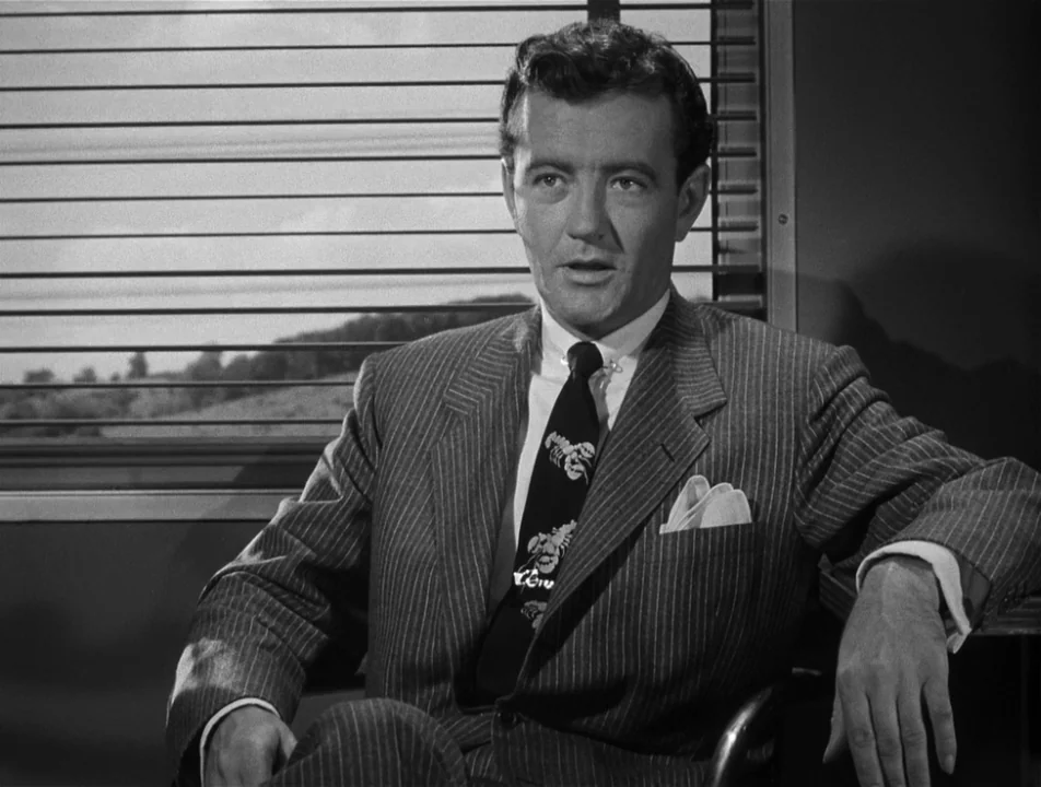 Strangers on a Train (1951) movie still: medium — A man sits in a train compartment with horizontal window blinds visible behind him. The man looks…