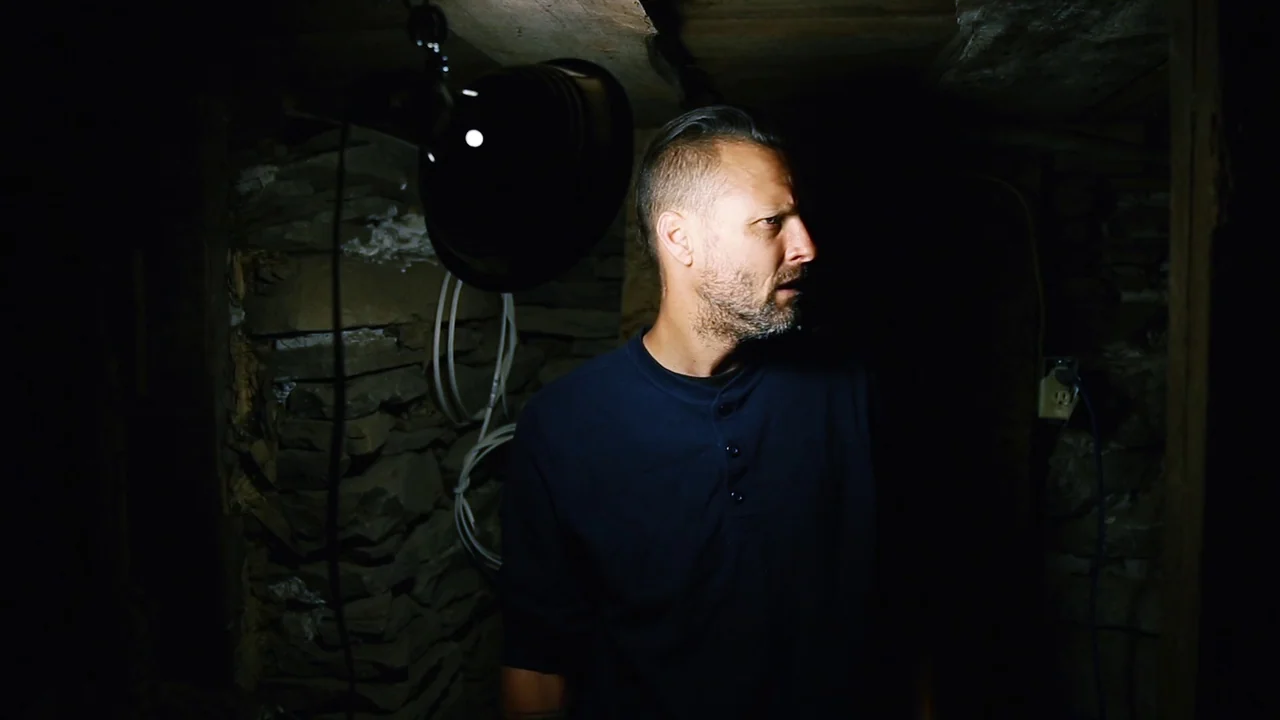 The Deeper You Dig (2019) movie still: medium — A dark, cramped basement with unfinished rough stone walls and exposed electrical wires. A man with…