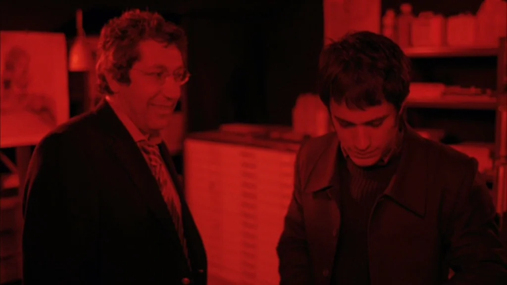 The Science Of Sleep (2006) movie still: medium — A darkroom with shelves holding bottles and equipment under red light. Two men are working…