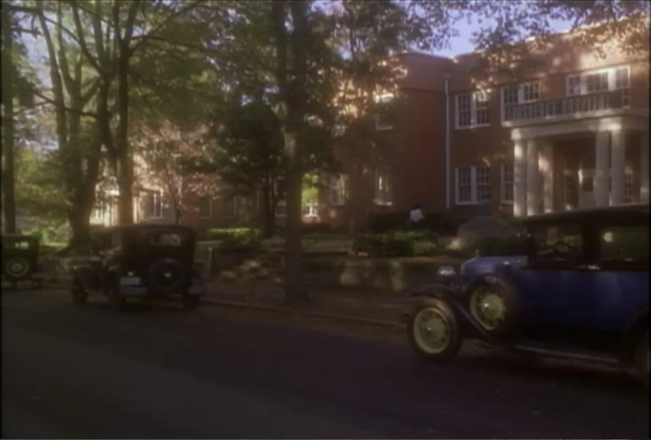 Miss Evers' Boys (1997) movie still: establishing — A brick hospital building stands behind a row of trees and parked vintage cars. The foreground…