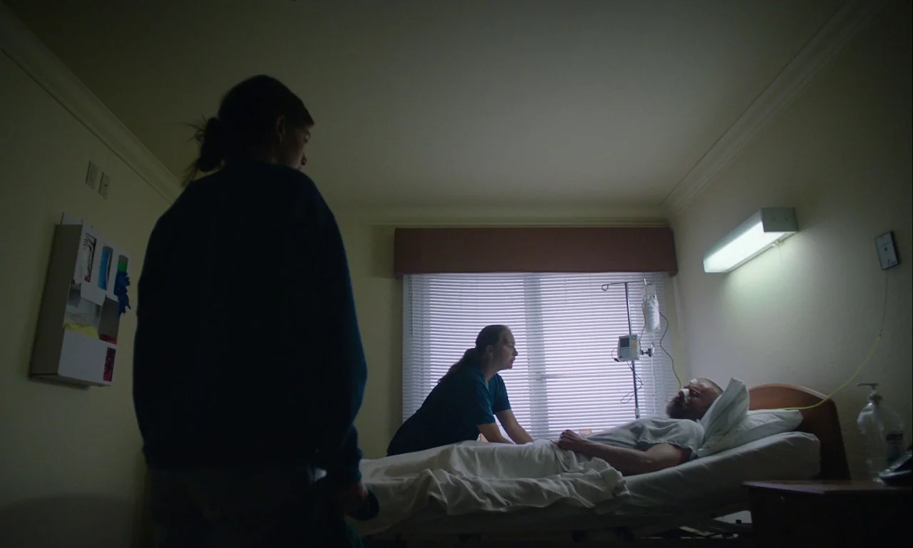 Mickey and the Bear (2019) movie still: wide — A hospital room has white walls, a patient bed, and a window with closed blinds. A nurse assists a…