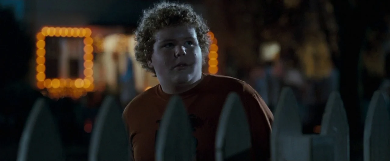 Trick ‘R Treat (2007) movie still: medium — A teenage boy stands in a residential neighborhood at night behind a wooden fence. Defocused orange…