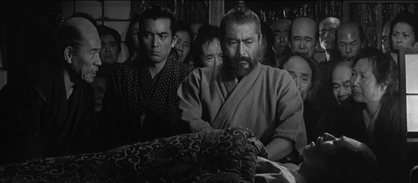 Red Beard (1965) movie still: medium — A group of people gathers inside a dimly lit traditional Japanese home at night. They stand closely together…