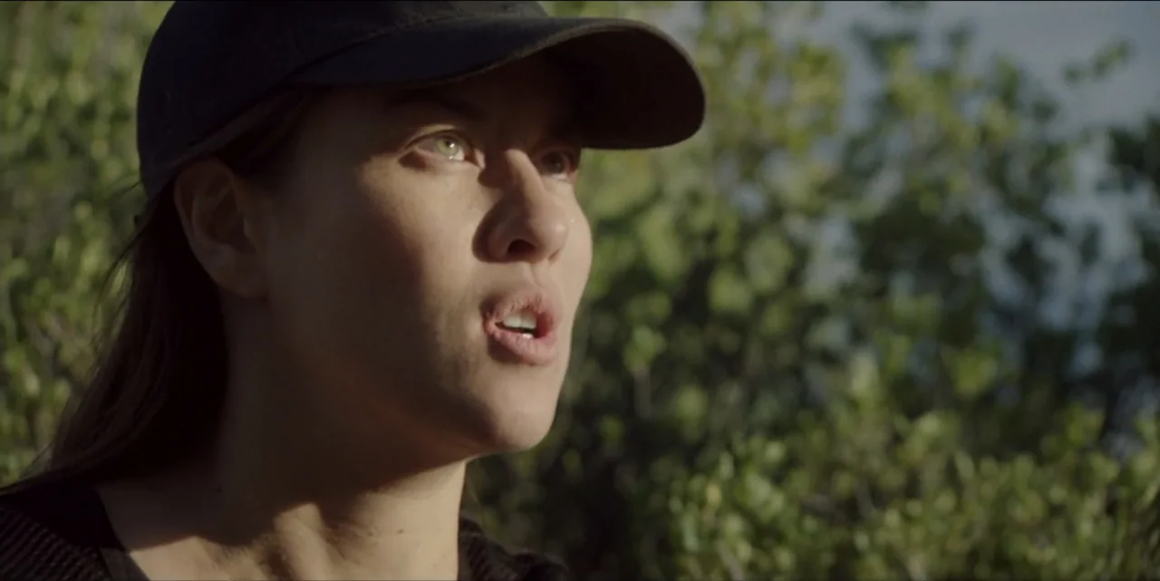 The Hunted (2024) movie still: closeup — A woman stands in front of dense green bushes during the late afternoon. She wears a baseball cap and has…