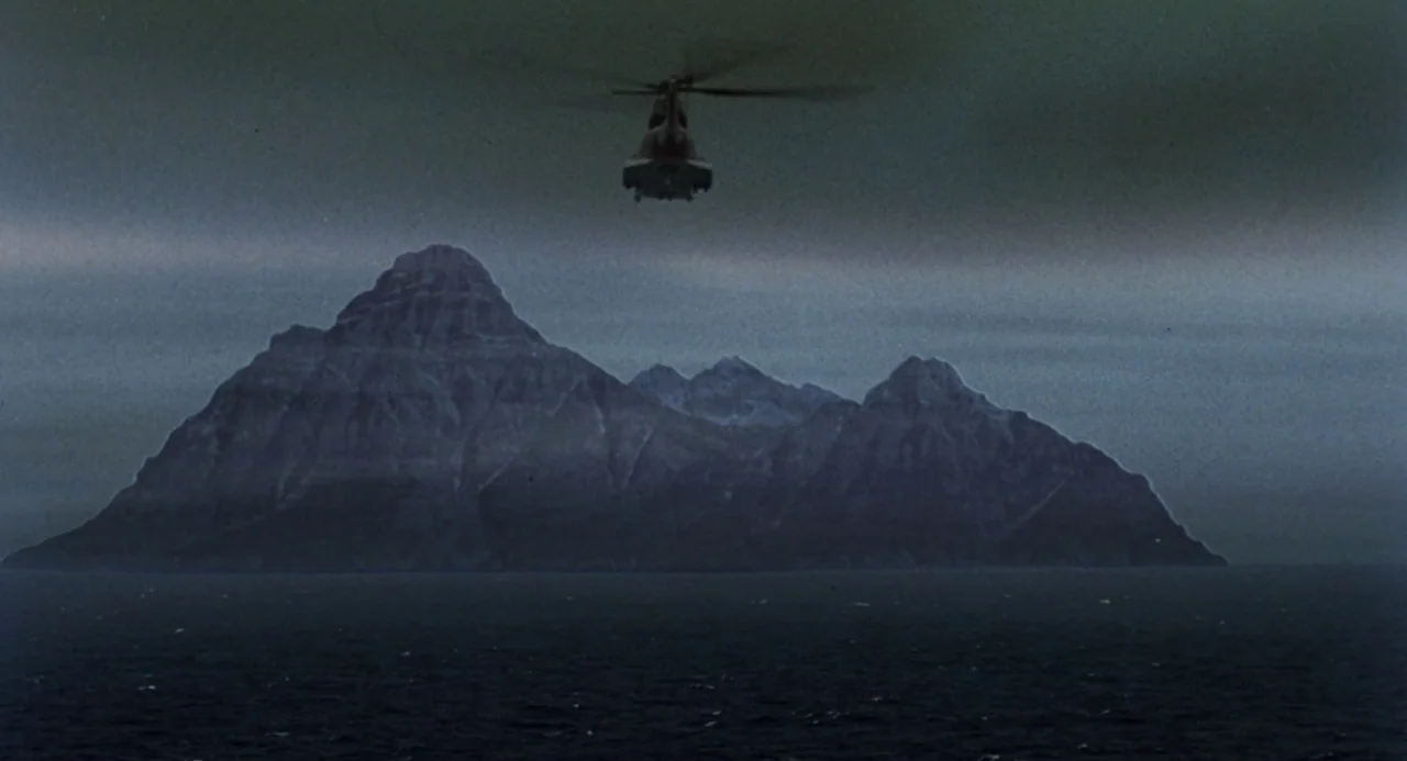 Godzilla vs. Mechagodzilla II (1993) movie still: establishing — A dark rocky island rises from a choppy grey sea under an overcast sky. A small…