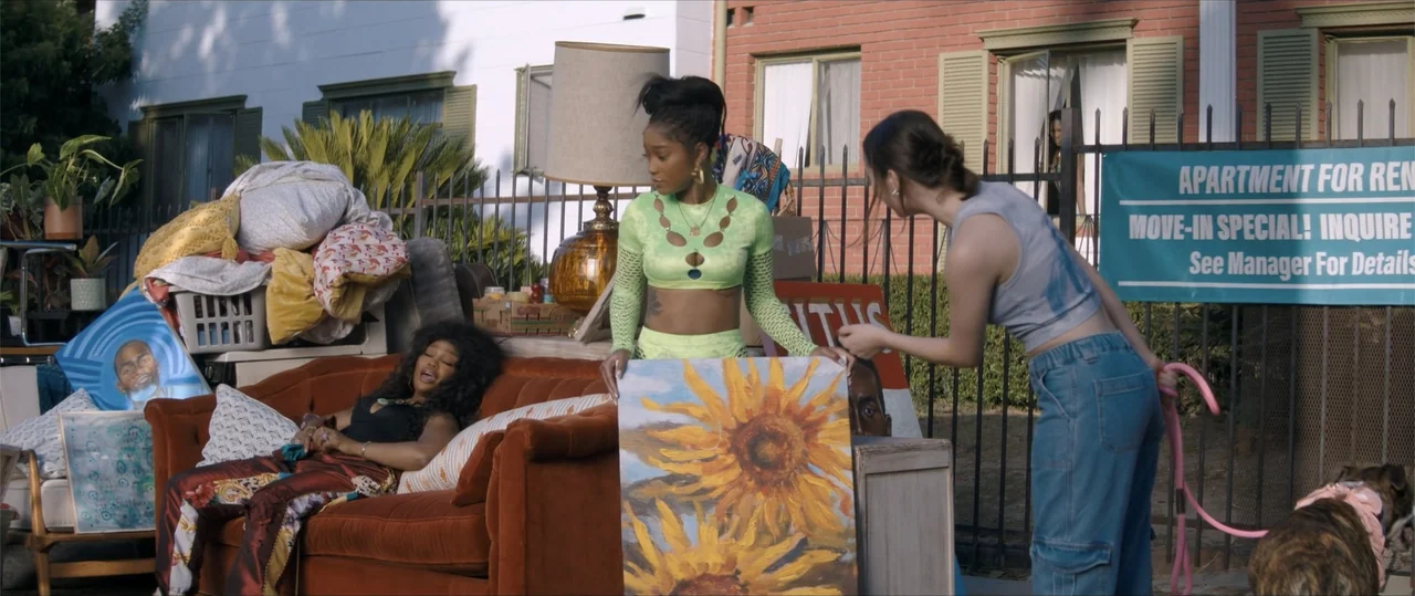 One of Them Days (2025) movie still: medium — Three young women arrange a variety of personal belongings and art on a city sidewalk. They are…
