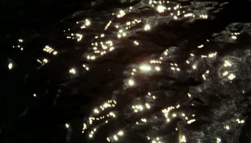 A Bay of Blood (1971) movie still: establishing — Dark water ripples with intense points of light reflecting off the moving surface. The frame is…