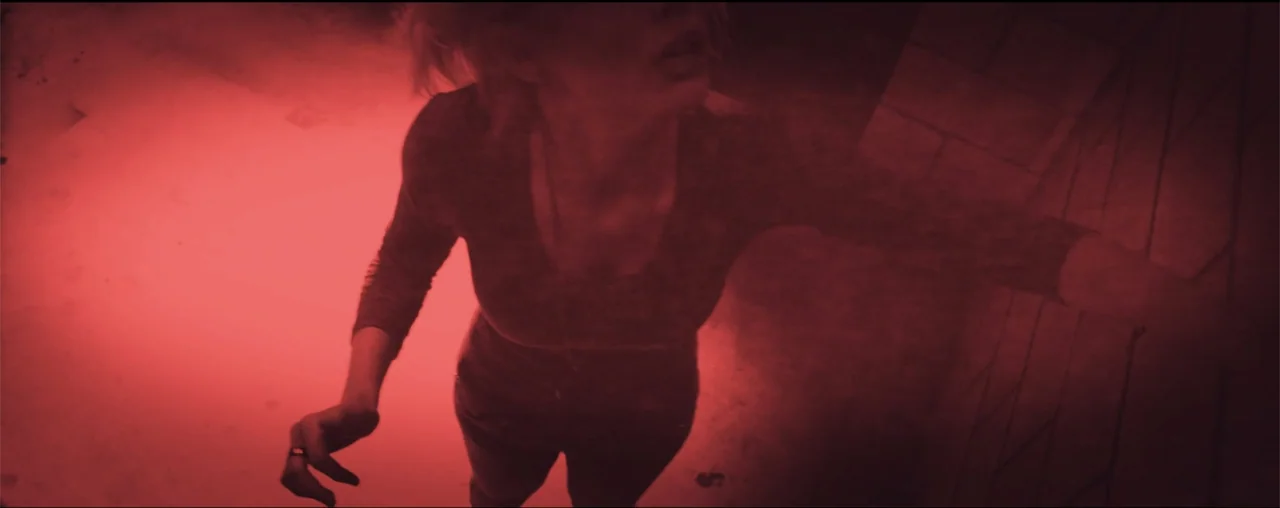 El Último Juego (2025) movie still: medium — A narrow interior corridor is illuminated by a harsh red light source. A woman stands in the center…