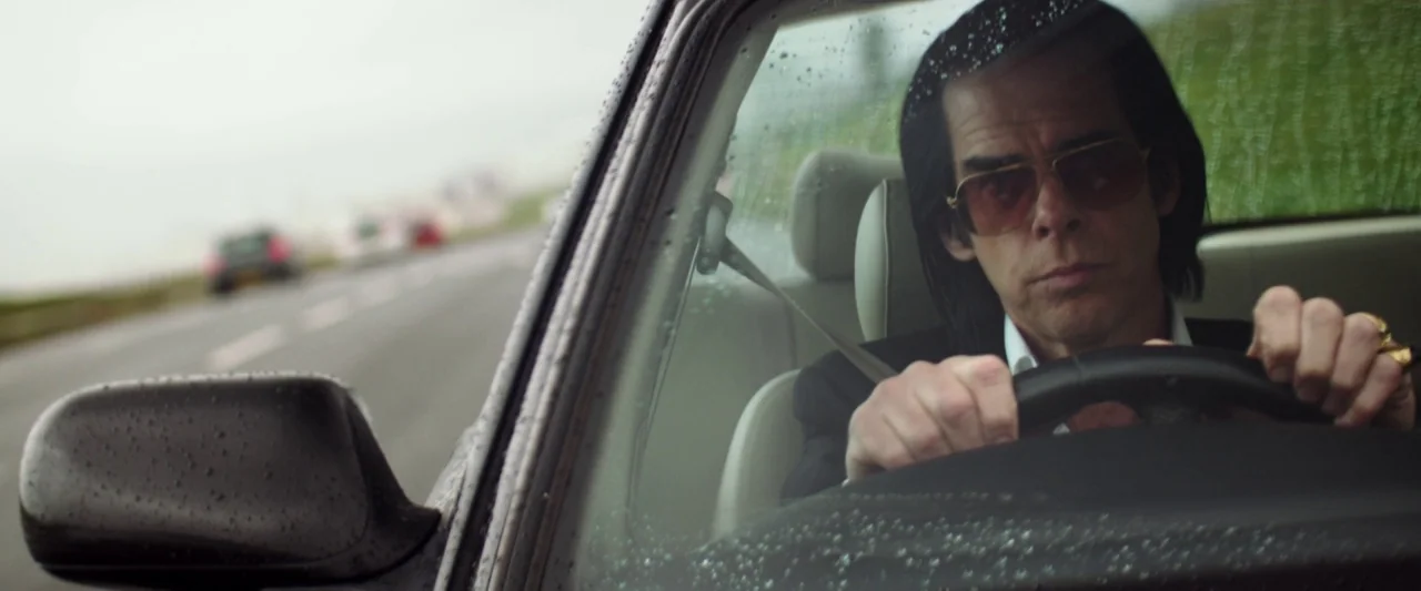 20,000 Days on Earth (2014) movie still: medium — A man drives a car on an overcast day with rain droplets on the glass. The camera frames him…