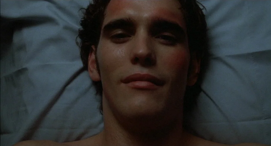 Drugstore Cowboy (1989) movie still: closeup — A man lies supine on a creased fabric surface in dim light. The subject is positioned horizontally in…